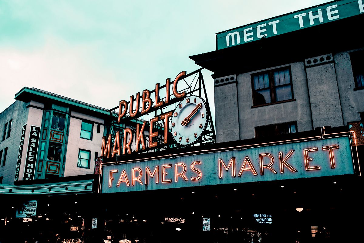The image shows a view of Pike Place Market with its famous clock and neon signs for "PUBLIC MARKET" and "FARMERS MARKET."