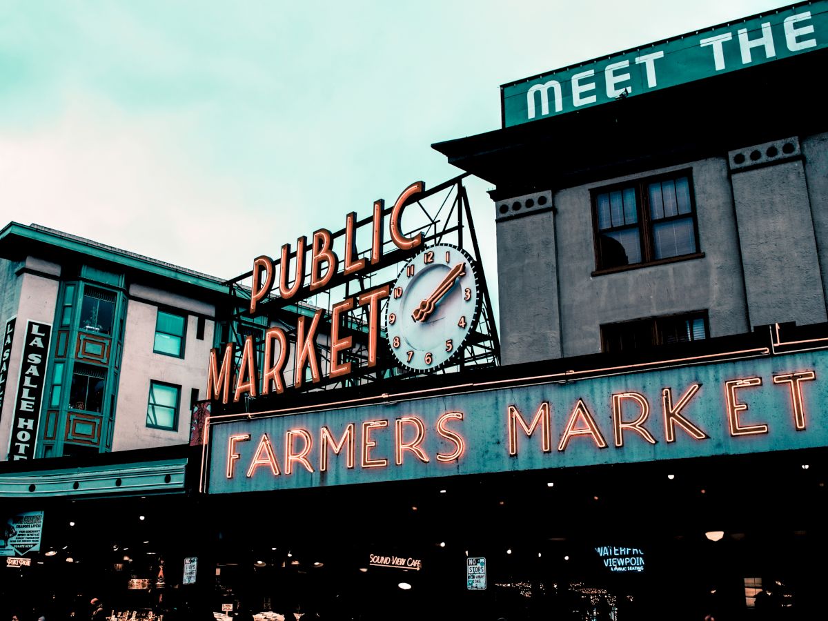 The image shows a view of Pike Place Market with its famous clock and neon signs for "PUBLIC MARKET" and "FARMERS MARKET."