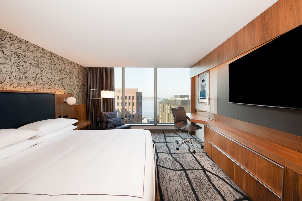 A modern hotel room with a large bed, wall-mounted TV, desk, and a window view of the city and water in the background.