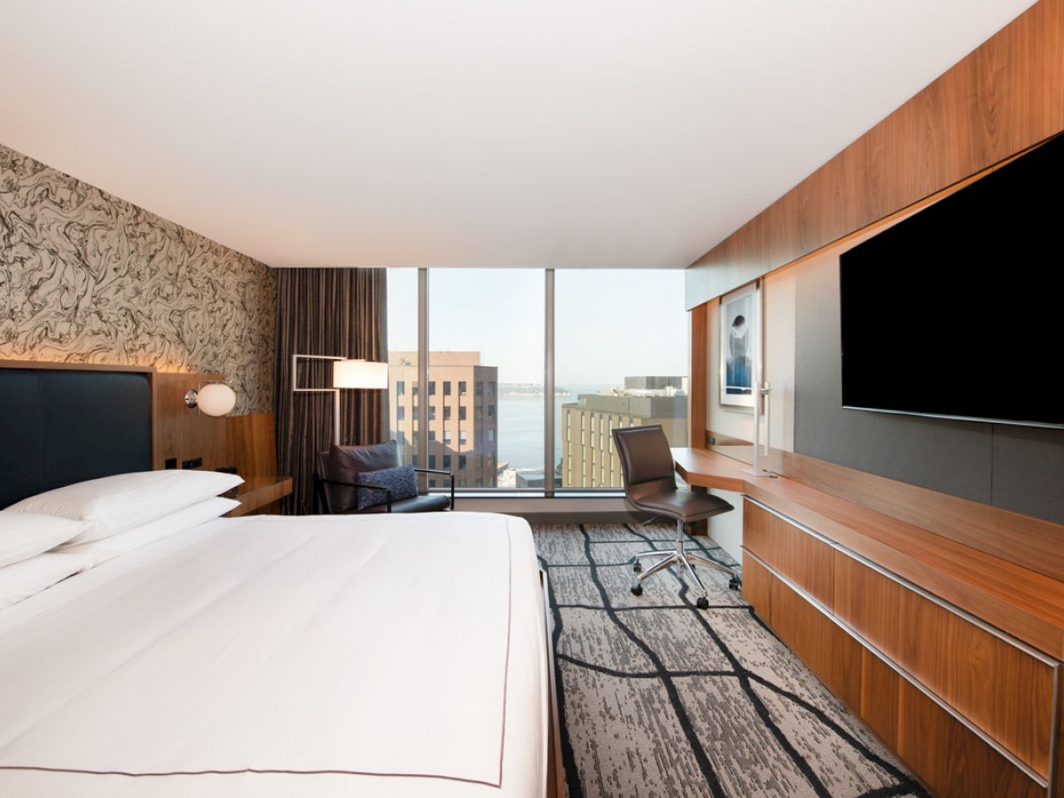 A modern hotel room with a large bed, wall-mounted TV, desk, and a window view of the city and water in the background.