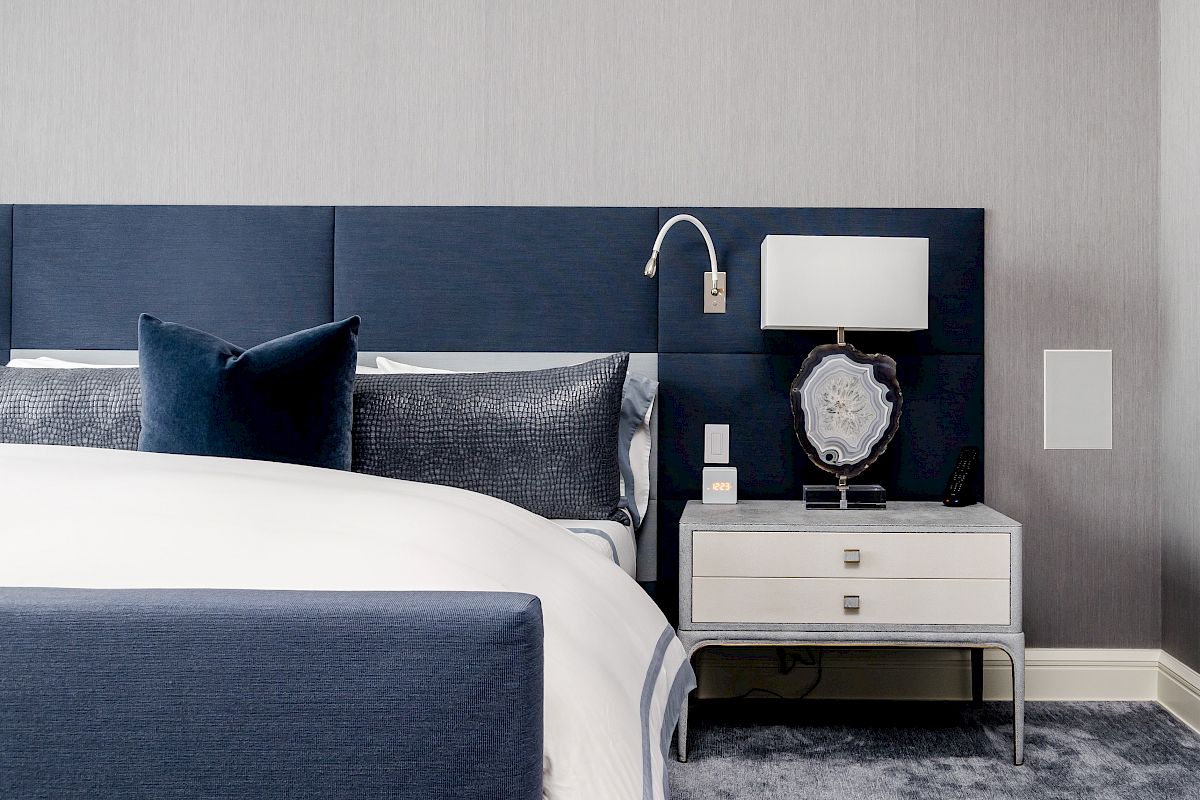 A modern bedroom with a navy blue headboard, white bed, gray carpet, and a nightstand with a lamp, decorative piece, and wall light.