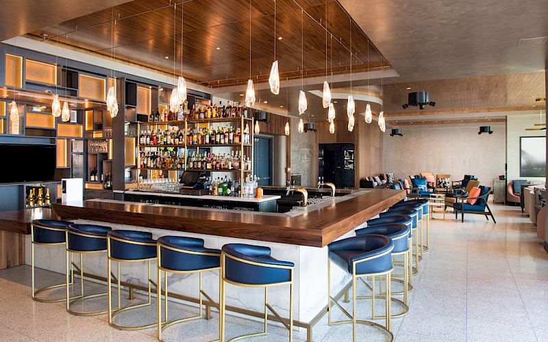 A modern bar with stylish blue seating, hanging lights, and a well-stocked back bar. The surrounding area features cozy seating.