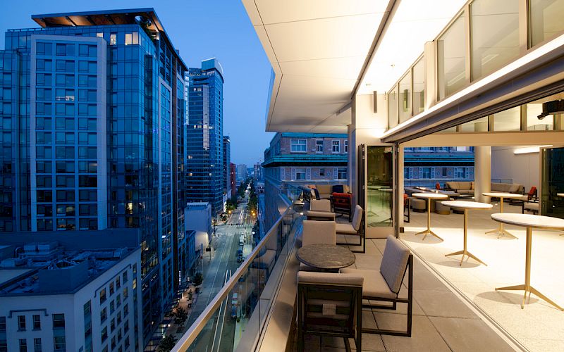 Rooftop terrace with seating overlooking a cityscape at twilight, adjacent to tall glass buildings and a street below.